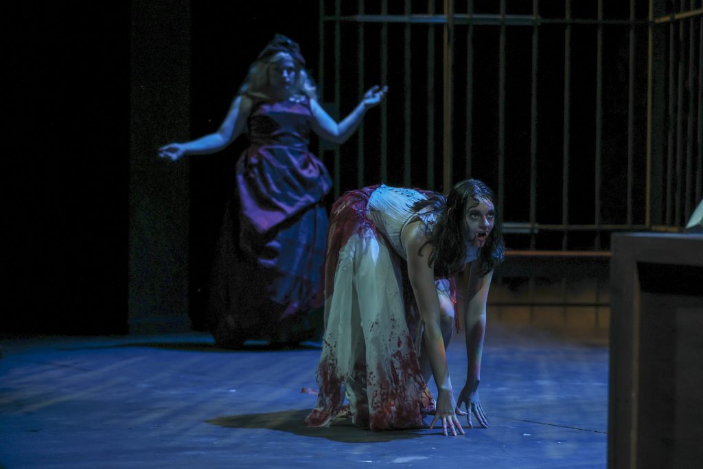 A bloodied woman crawls across the stage under blue lighting as another woman stands behind her in Pierce College Theatre's Dracula.