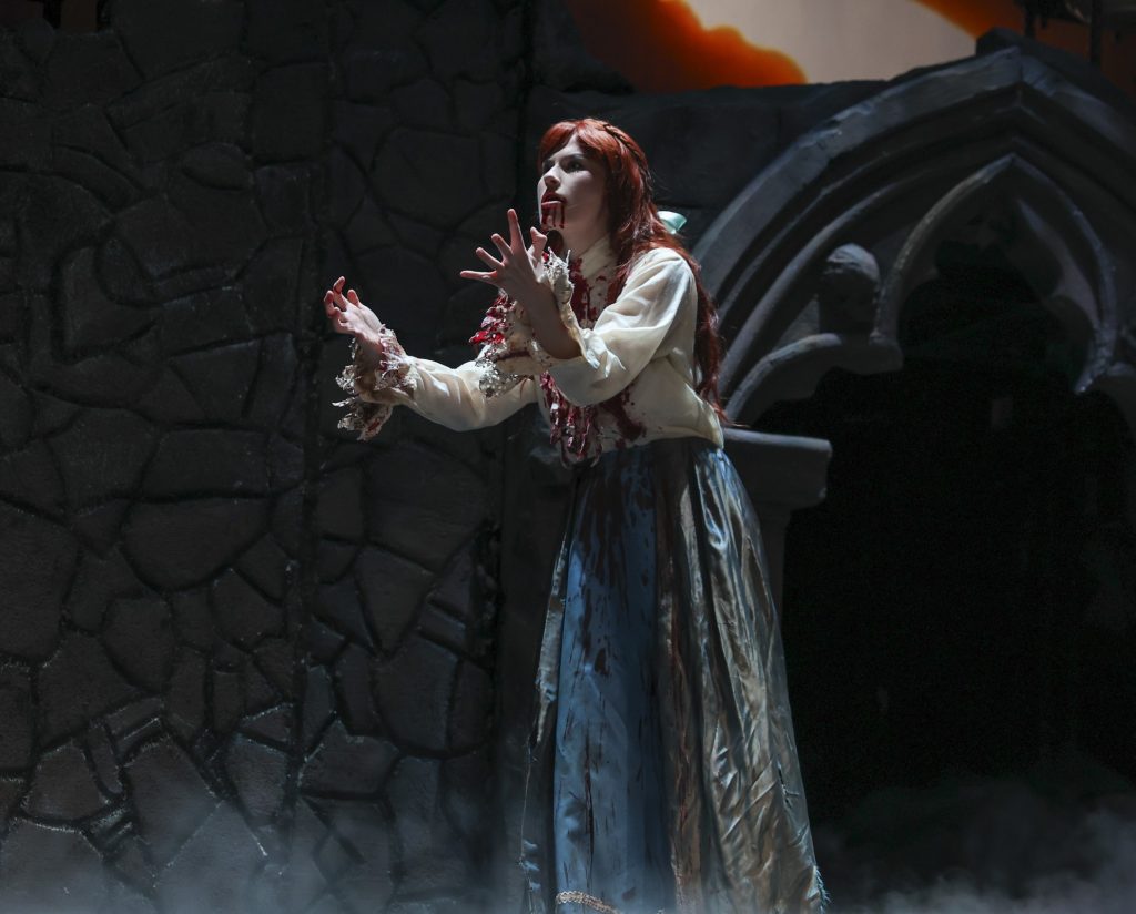 A bloodied red-haired woman reaches outward in anguish against a gothic stone backdrop during Pierce College Theatre's Dracula.
