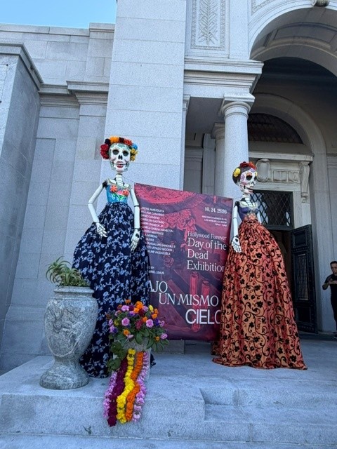 IMAGE_1 Two colorful Día de los Muertos skeleton sculptures in traditional dresses stand outside Hollywood Forever's Cathedral Mausoleum beside the Bajo un Mismo Cielo exhibition banner.