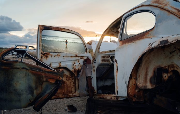 IMAGE_1 Katherine Flynn installation featuring rusted Volkswagen car parts and a photograph of a woman standing in the ocean, blending memory, travel and found materials.
