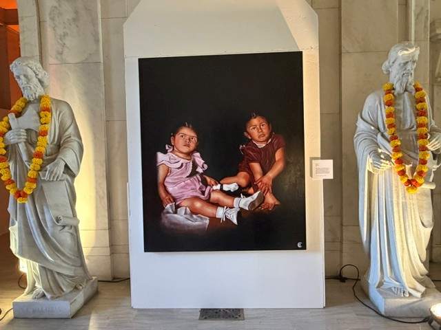 Painting of two young girls displayed between marble statues adorned with marigold garlands at Hollywood Forever's Día de los Muertos art exhibition.