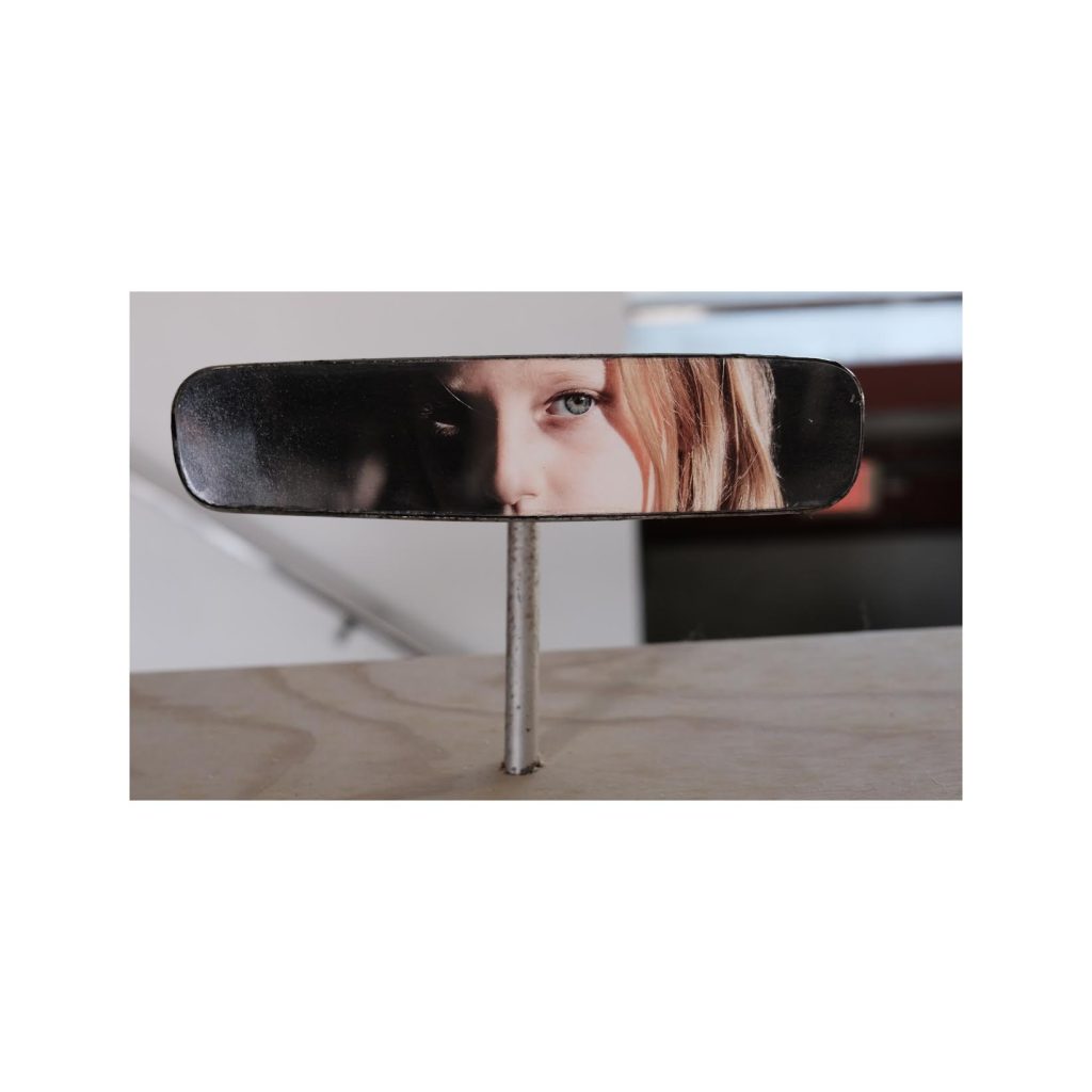 Katherine Flynn artwork featuring a rearview mirror with a close-up photograph of a woman’s eye reflected across its surface.