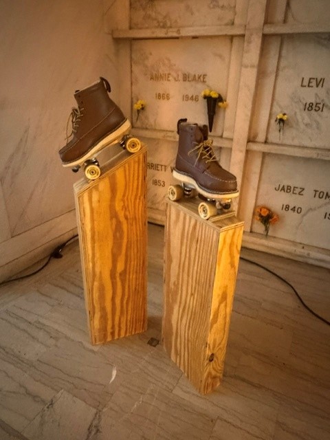 Two roller skates displayed on wooden pedestals in front of marble memorial niches inside Hollywood Forever's Cathedral Mausoleum.