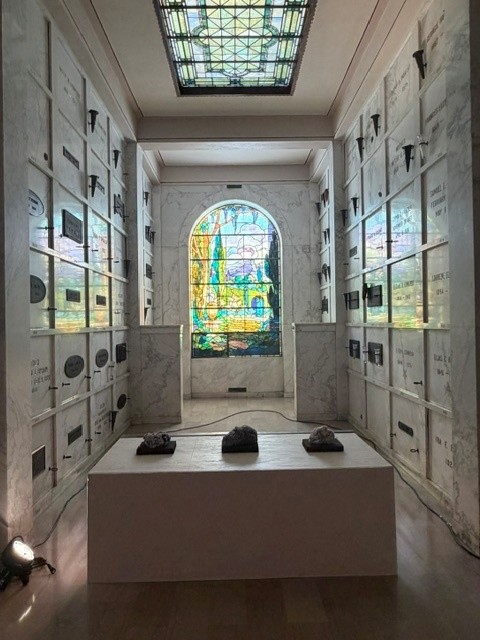 Interior of Hollywood Forever's Cathedral Mausoleum featuring marble memorial walls, a stained-glass window and a contemporary art installation displayed on a white pedestal.