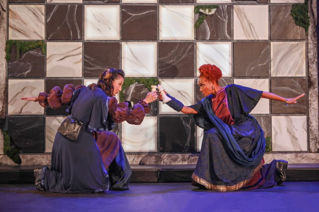 Two actors in Crone Creed kneel onstage facing each other with goblet-like props against a large checkerboard set.