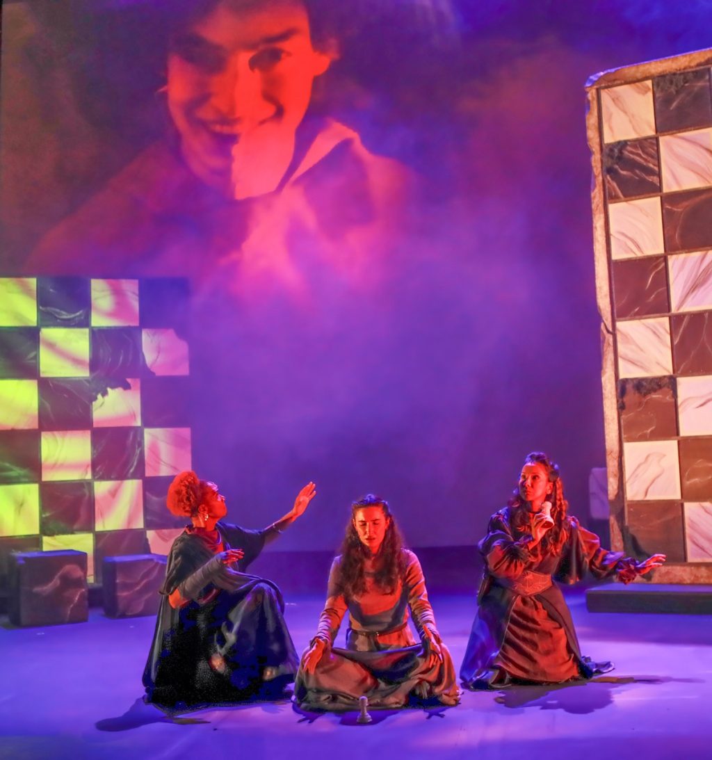 Three actors in Crone Creed perform under vivid purple and red lighting with a large projected face above the stage.