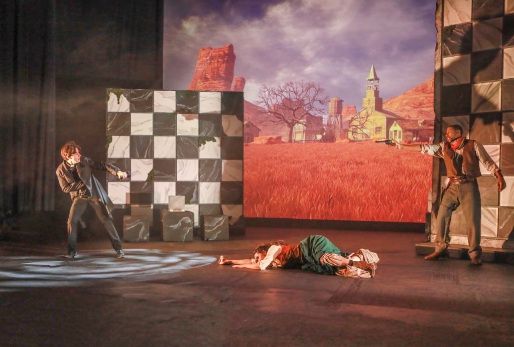 Dramatic confrontation in Crone Creed with two armed actors and another character lying onstage before a projected landscape.