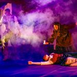 Dramatic scene from Crone Creed at El Portal Theatre with actors surrounded by theatrical smoke and purple lighting.
