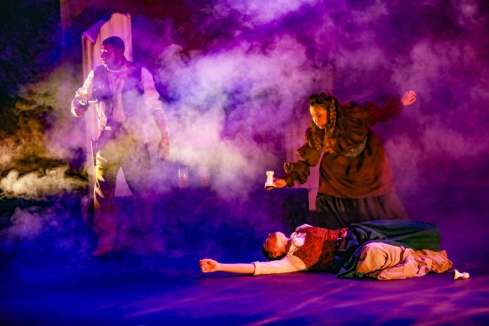 Dramatic scene from Crone Creed at El Portal Theatre with actors surrounded by theatrical smoke and purple lighting.
