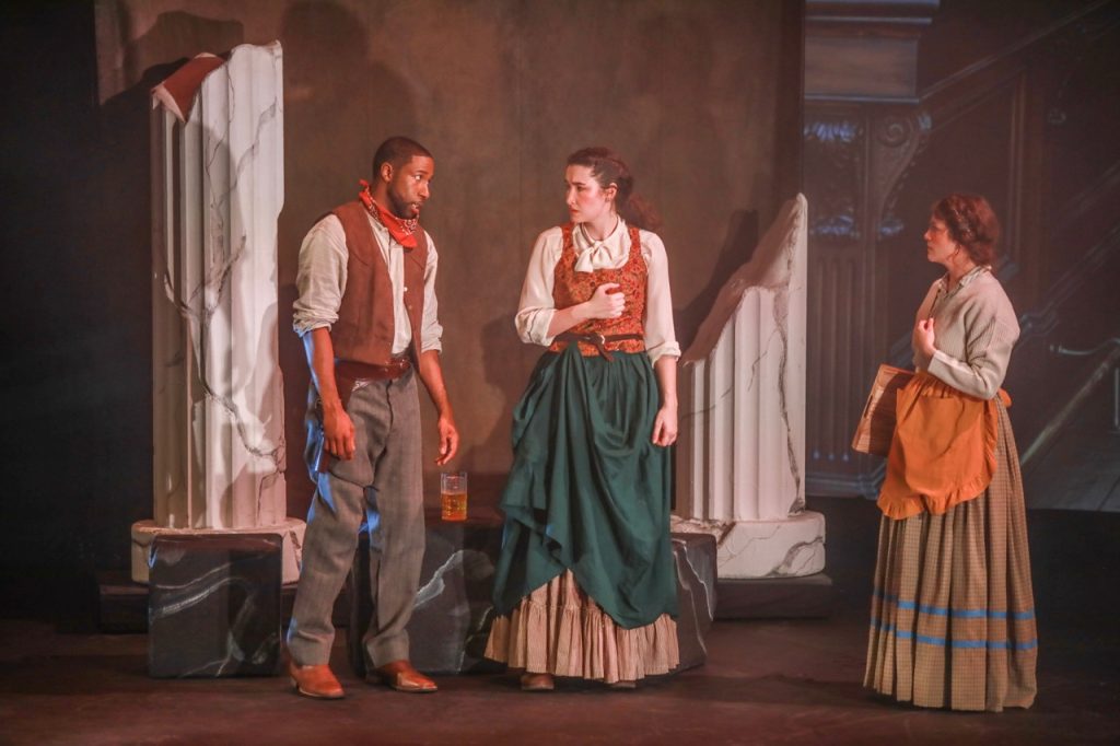 Three actors perform a period scene in Crone Creed amid the production’s stylized set and projected scenery.