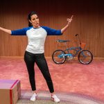 Denise Fennell performs Lessons Learned: The Time of My Life onstage with a bicycle and simple set pieces behind her.