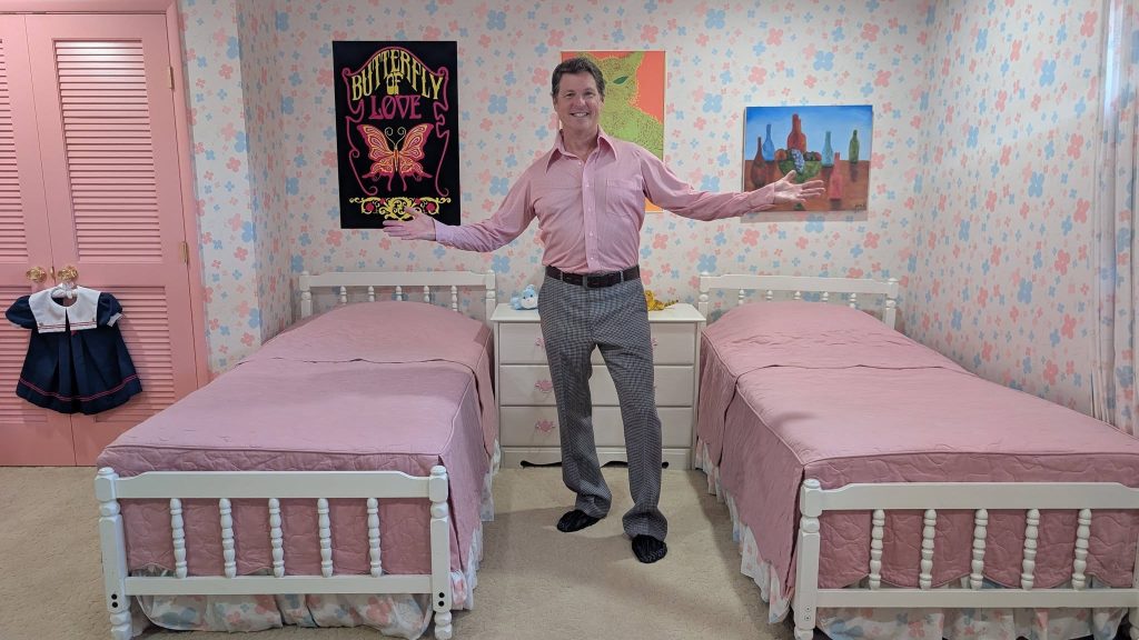 Jack Witt poses between two pink twin beds in the recreated Brady girls' bedroom, featuring floral wallpaper and 1970s decor.