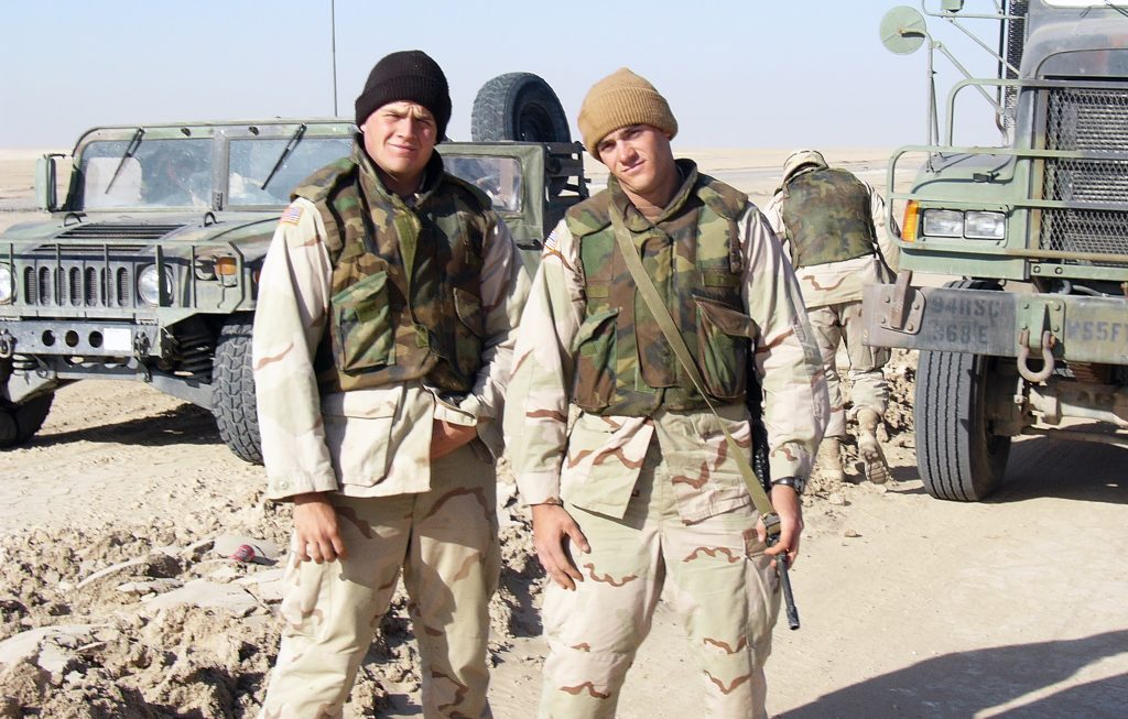 Army veteran and RustRiders musician Ryan Curtis with fellow soldier Rob in military uniform beside U.S. Army vehicles during deployment.